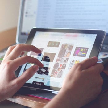 A pair of hands scrolling through e-commerce products on a touch-screen tablet with a laptop also open in the background.