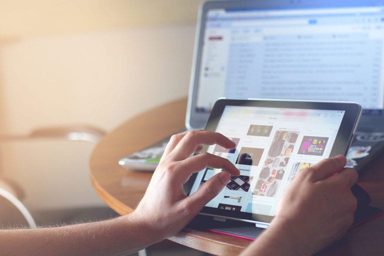 A pair of hands scrolling through e-commerce products on a touch-screen tablet with a laptop also open in the background.