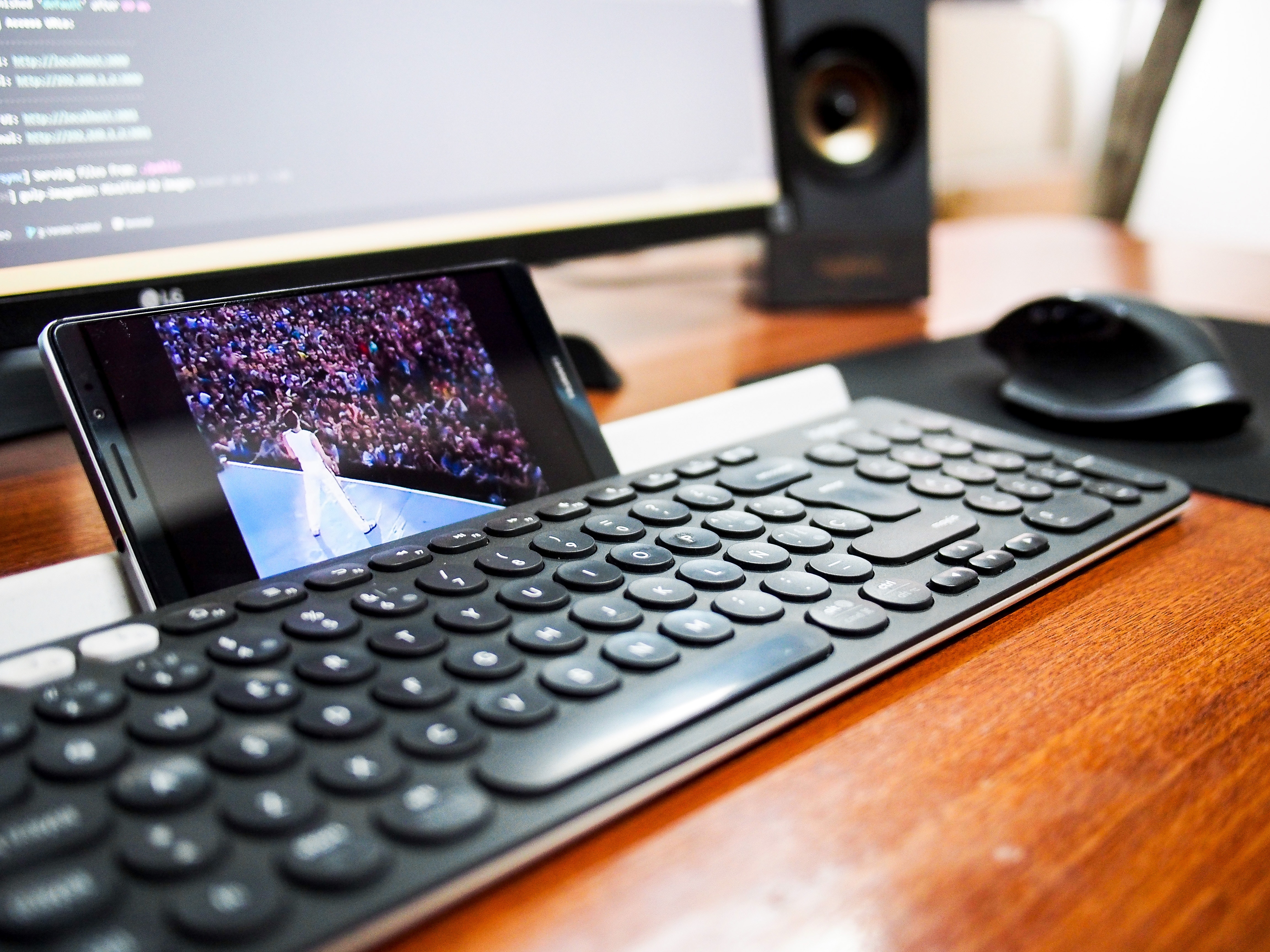 A cell phone propped between a computer keyboard and monitor, playing a video.