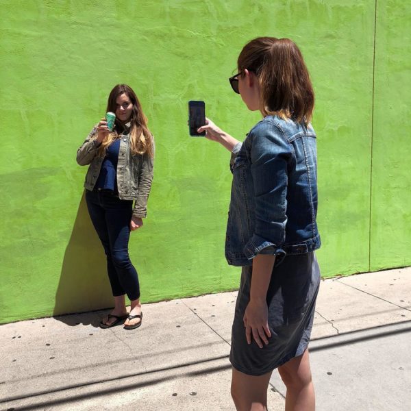 Young woman takes a photo of another woman drinking La Croix by a green wall.