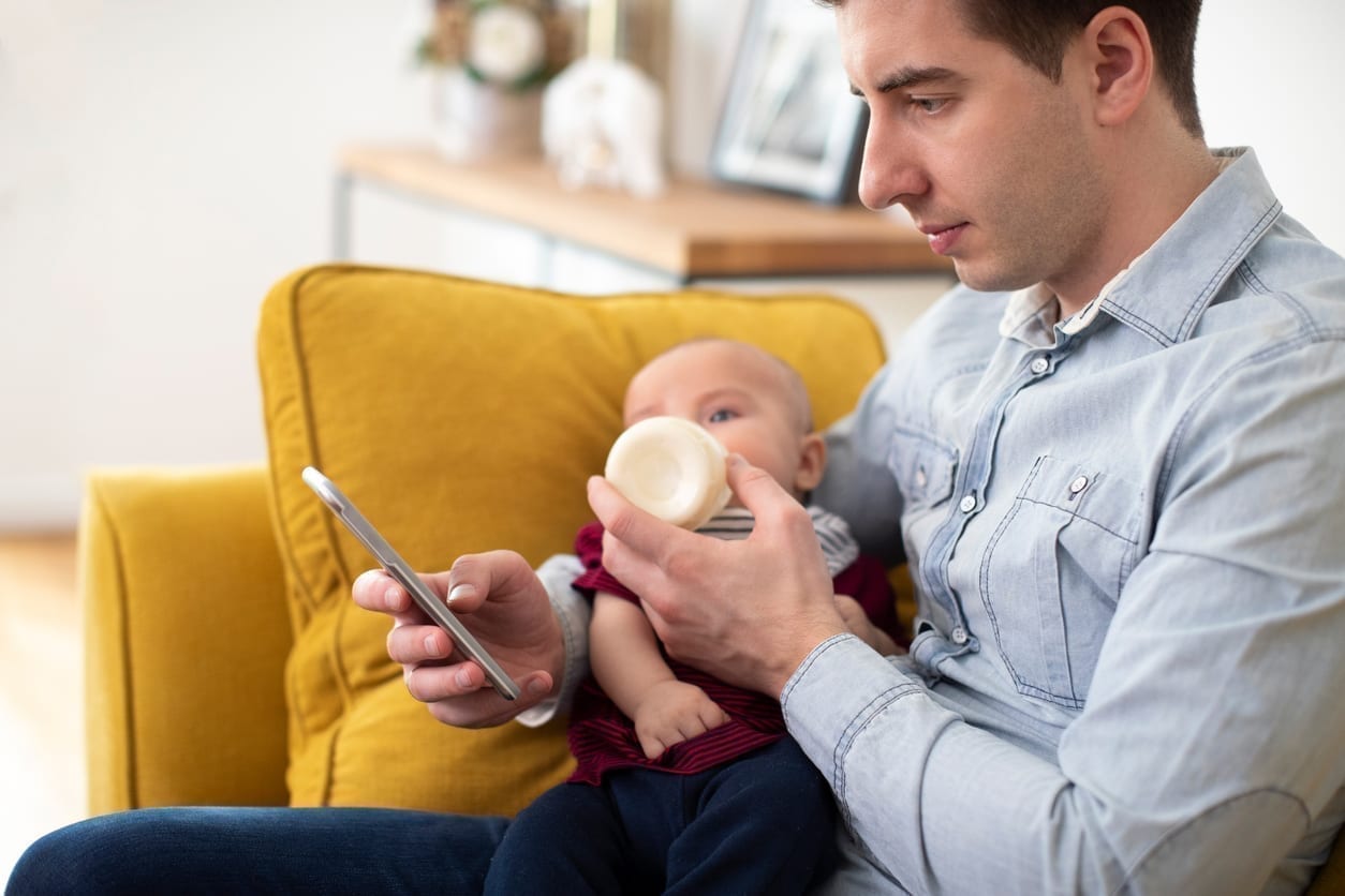 A father holds a bottle and a smartphone to work from home with a baby.