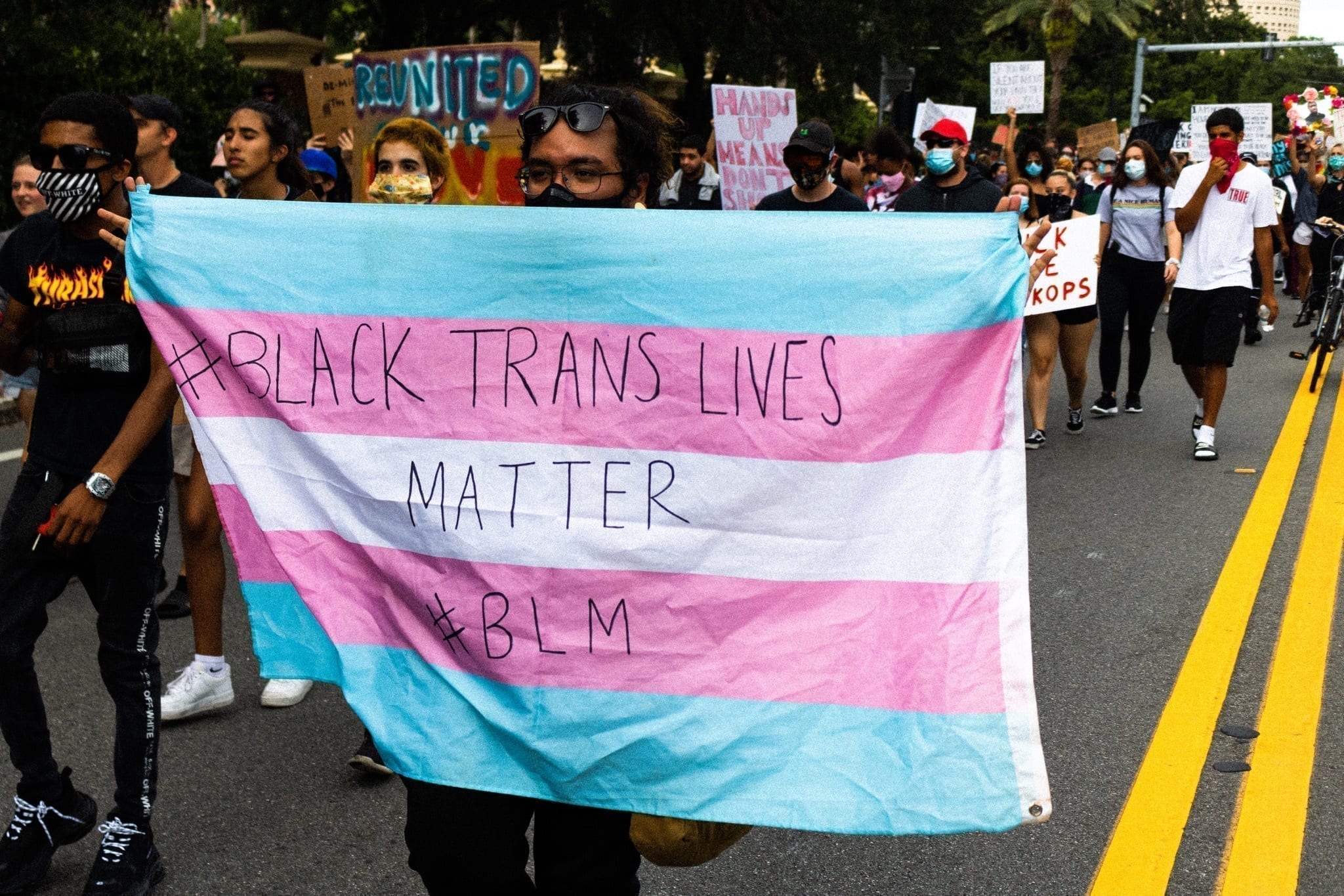 In a protest, a person holds a blue, pink, and white flag that reads "Black Trans Lives Matter BLM"