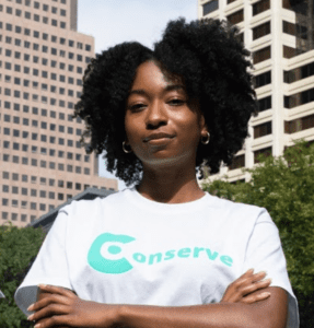 Girl wearing a natural twist out smiling at the camera with arms crossed confidently she's wearing a white Conserve Innovations t-shirt.