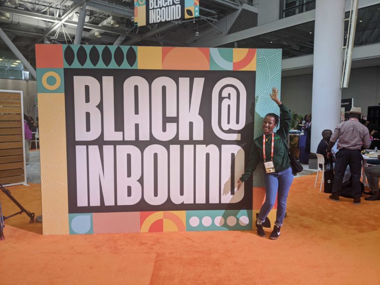 Our Business Development Director, Irene Muchai at the Hubspot Inbound Conference, standing next to a sign that reads "Black @ Inbound"