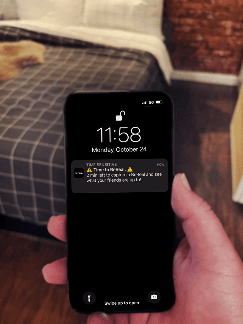The BeReal notification "Time to BeReal: 2 min left to capture a BeReal and see what your friends are up to!" pops up on a black iPhone in someone's hand. Behind the phone is a wood floor and a bed with a windowpane-patterned comforter.