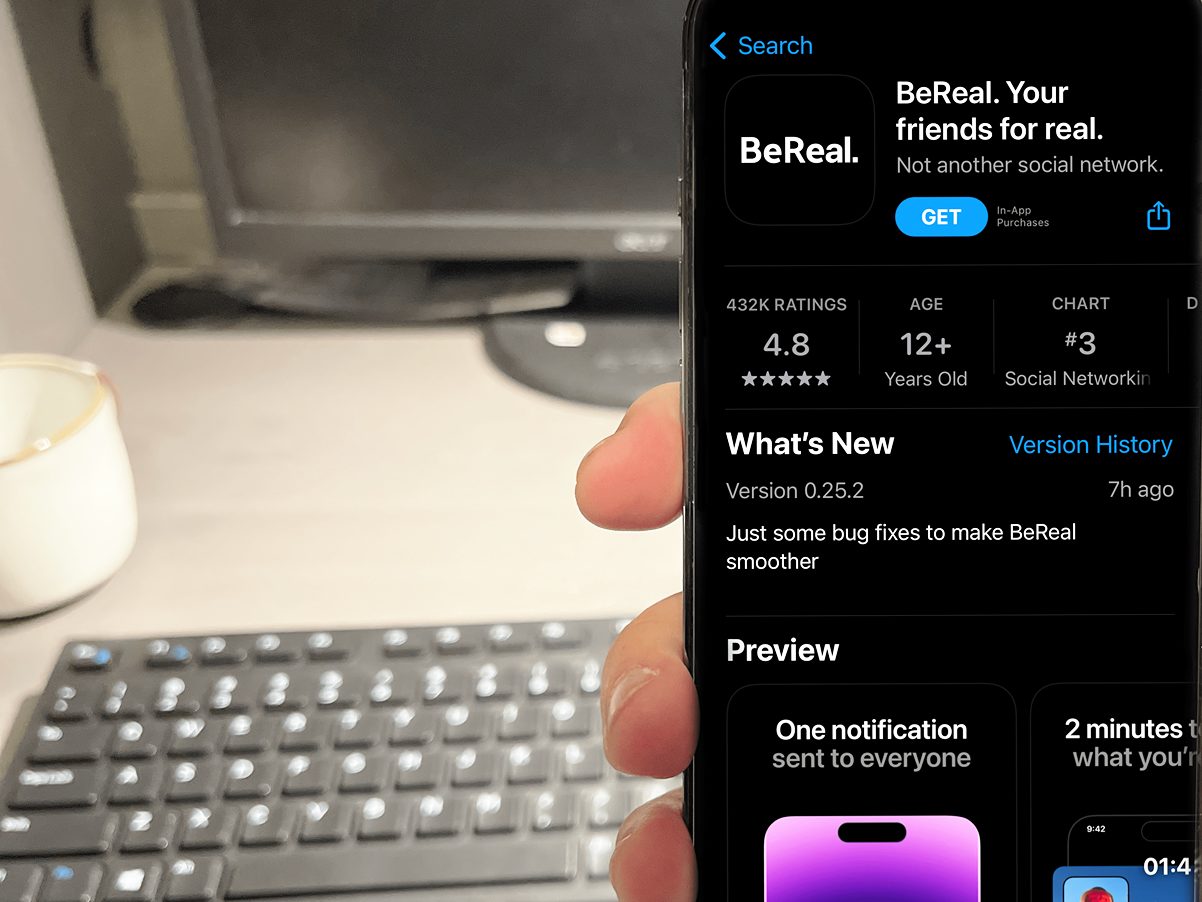 The BeReal app displayed in the app store on an iPhone, held in front of a computer keyboard on a desk.