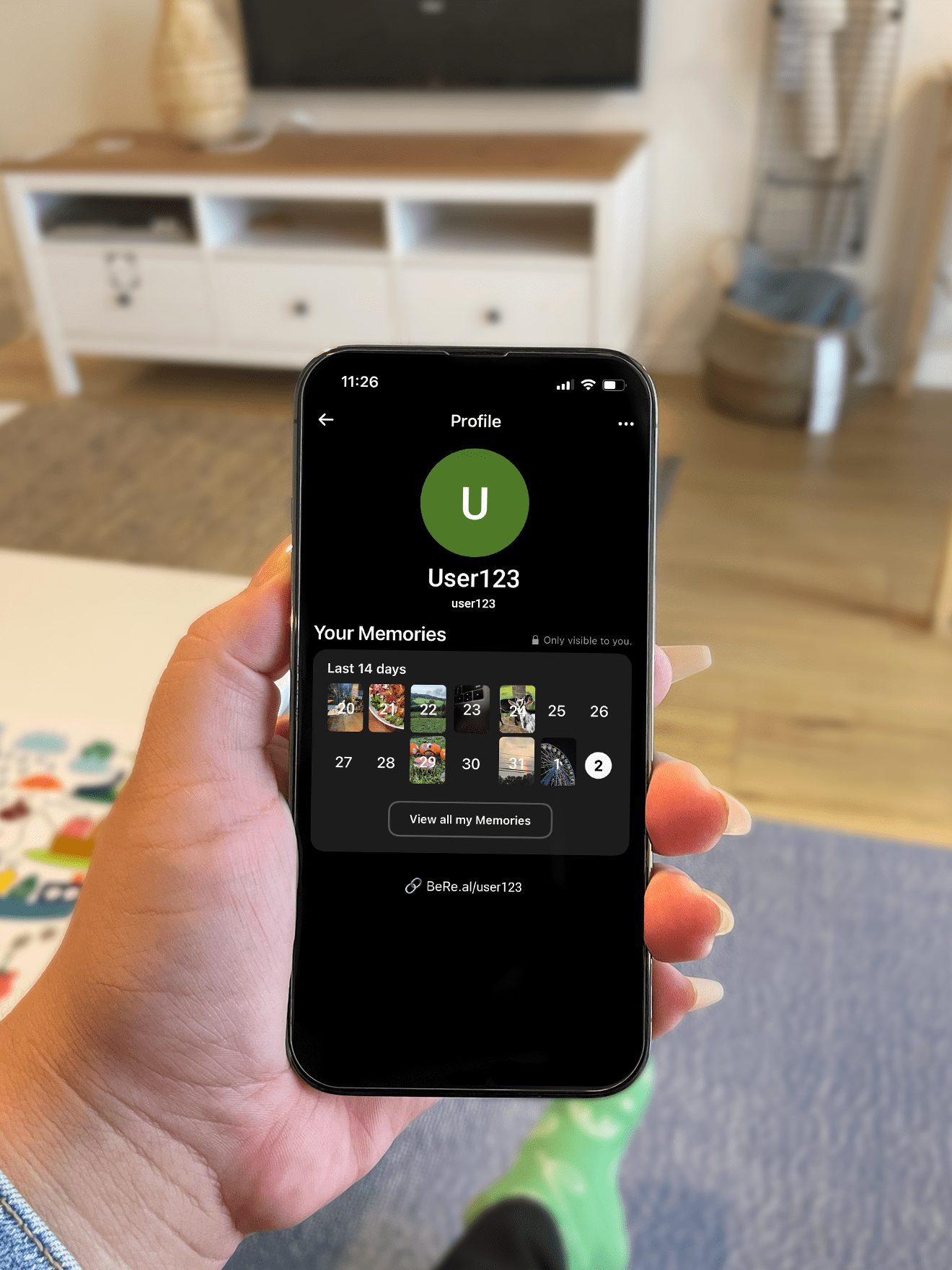 A sample user profile on the BeReal app ("User123") is displayed on an iPhone held in a person's hand. Behind the iPhone is a cozy living room.