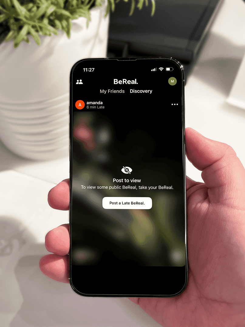 A phone displaying the "Post a Late BeReal" screen, held in front of a desk with a green plant on it.