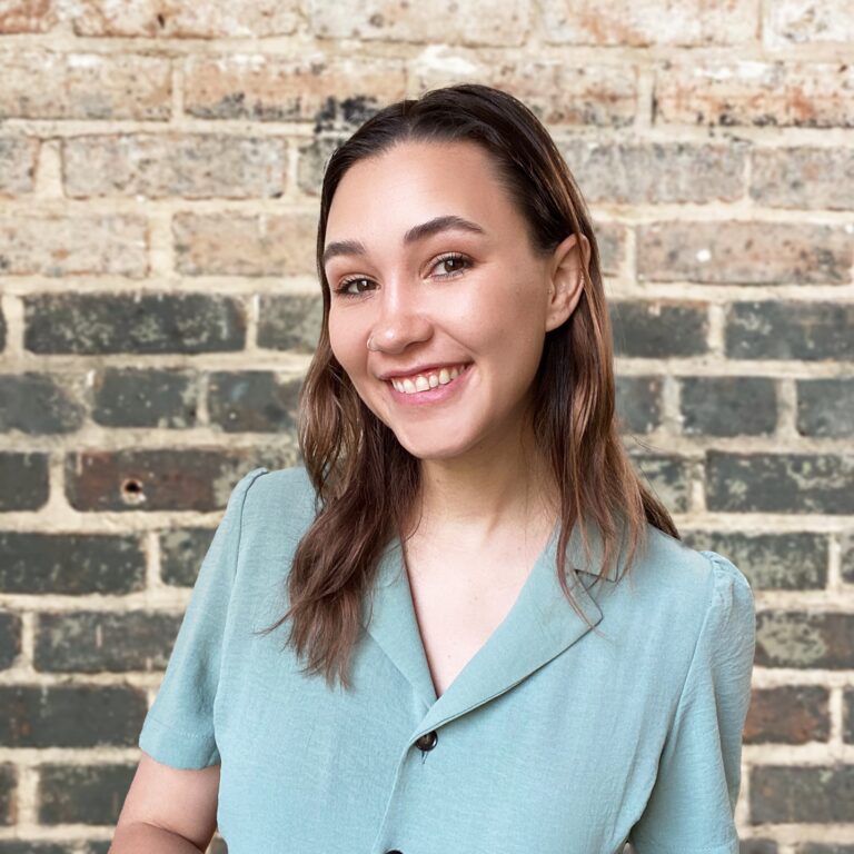 Meara McNitt, wearing a blue dress with a black belt and standing in front of a brick wall, smiling at the camera.