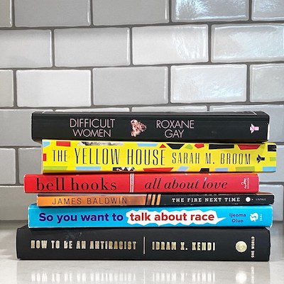 A stack of several of our DEI book club books, including Difficult Women, The Yellow House, All About Love, The Fire Next Time, So You Want to Talk About Race, and How to Be an Antiracist