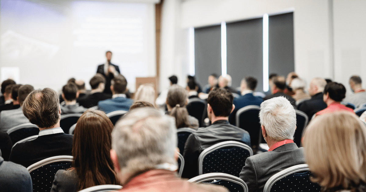 A crowd of people attending one of the best manufacturing conferences of the year listens to a speaker